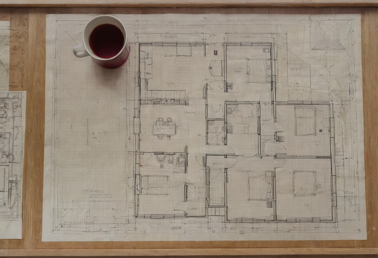 An old ink floor plan of a house on linen drawing paper mounted on a timber board, with a mug of tea standing on it and the edge of a second drawing beneath.