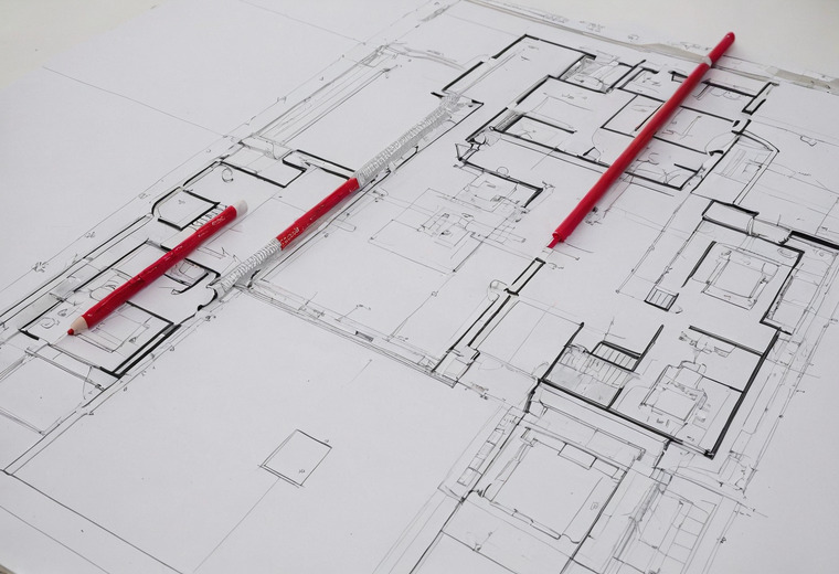 A large printed floor plan seen at an angle with two red pencils and a white ruler lying across it.